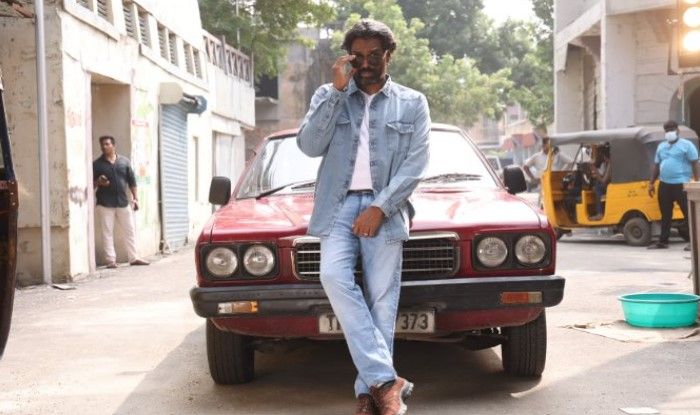 Image of Romesh Ranganathan on the phone leaning against a red car on an urban street.